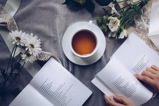 An overhead view of a table on which sit a couple of books and a cup of coffee. A pair of hands holds one book open, as if reading from it.