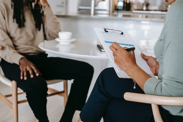 Two people sit at a table with coffee before them. We can see their torsos and lower bodies. One has a clipboard and is taking notes.