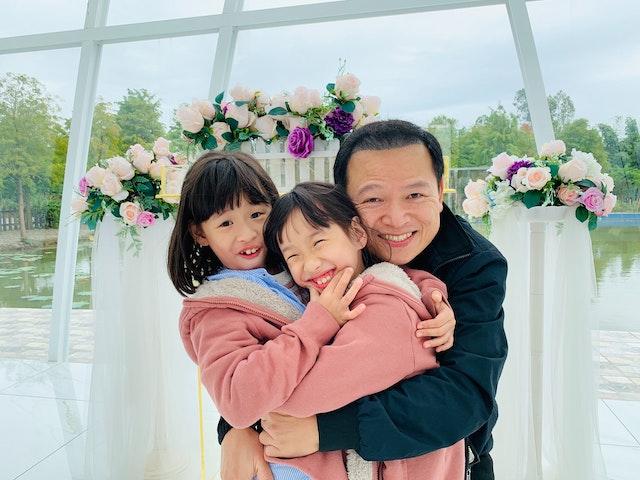 A man hugs his two daughters in front of a display of roses.