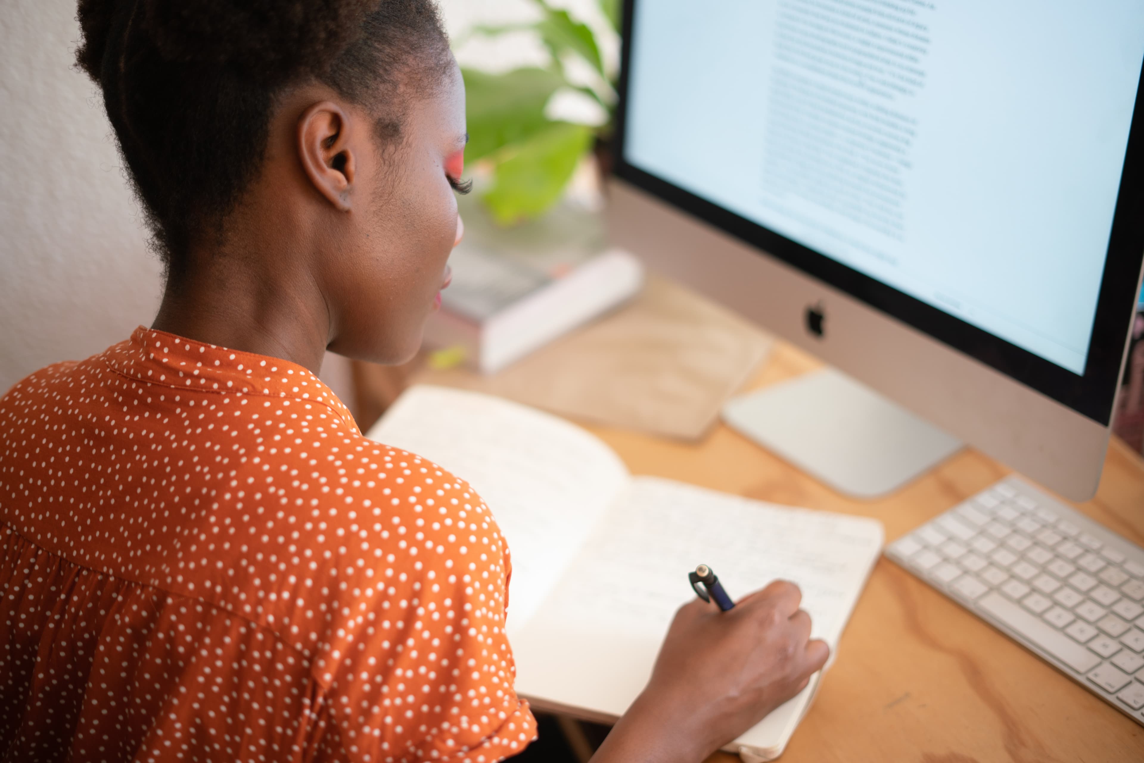 Young, brightly dressed woman seated at a computer writes in a notebook.